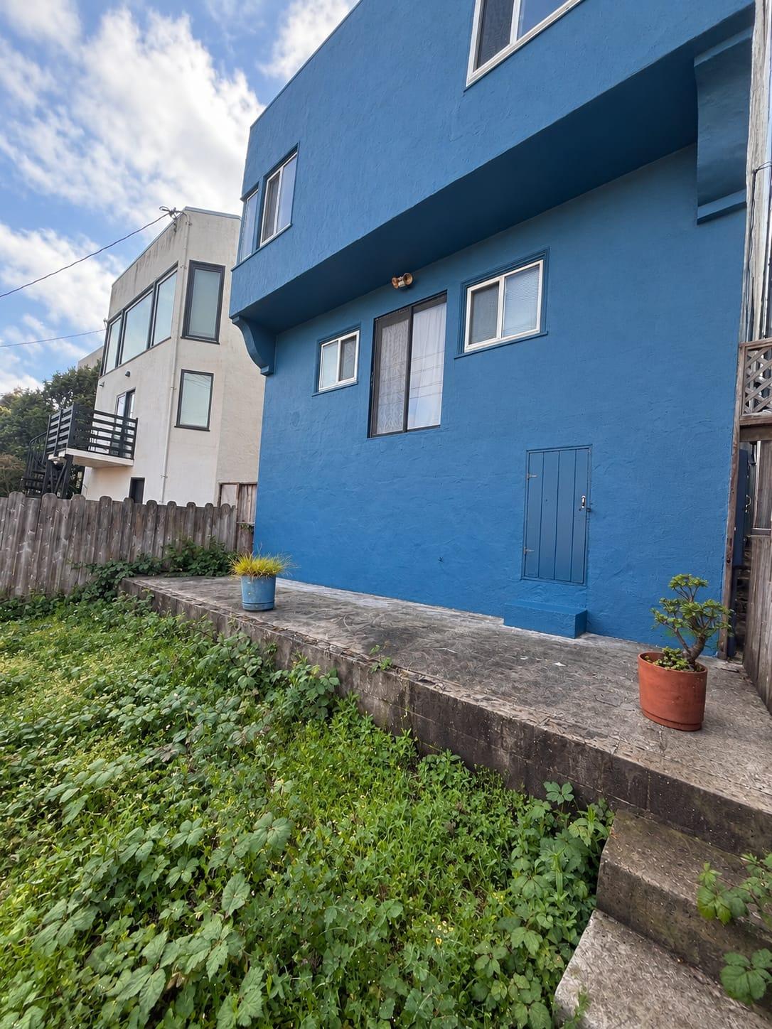 Back of a blue stucco Bay Area home with a bare wall over a raised concrete patio and an overgrown yard, before Gadget Construction built a deck