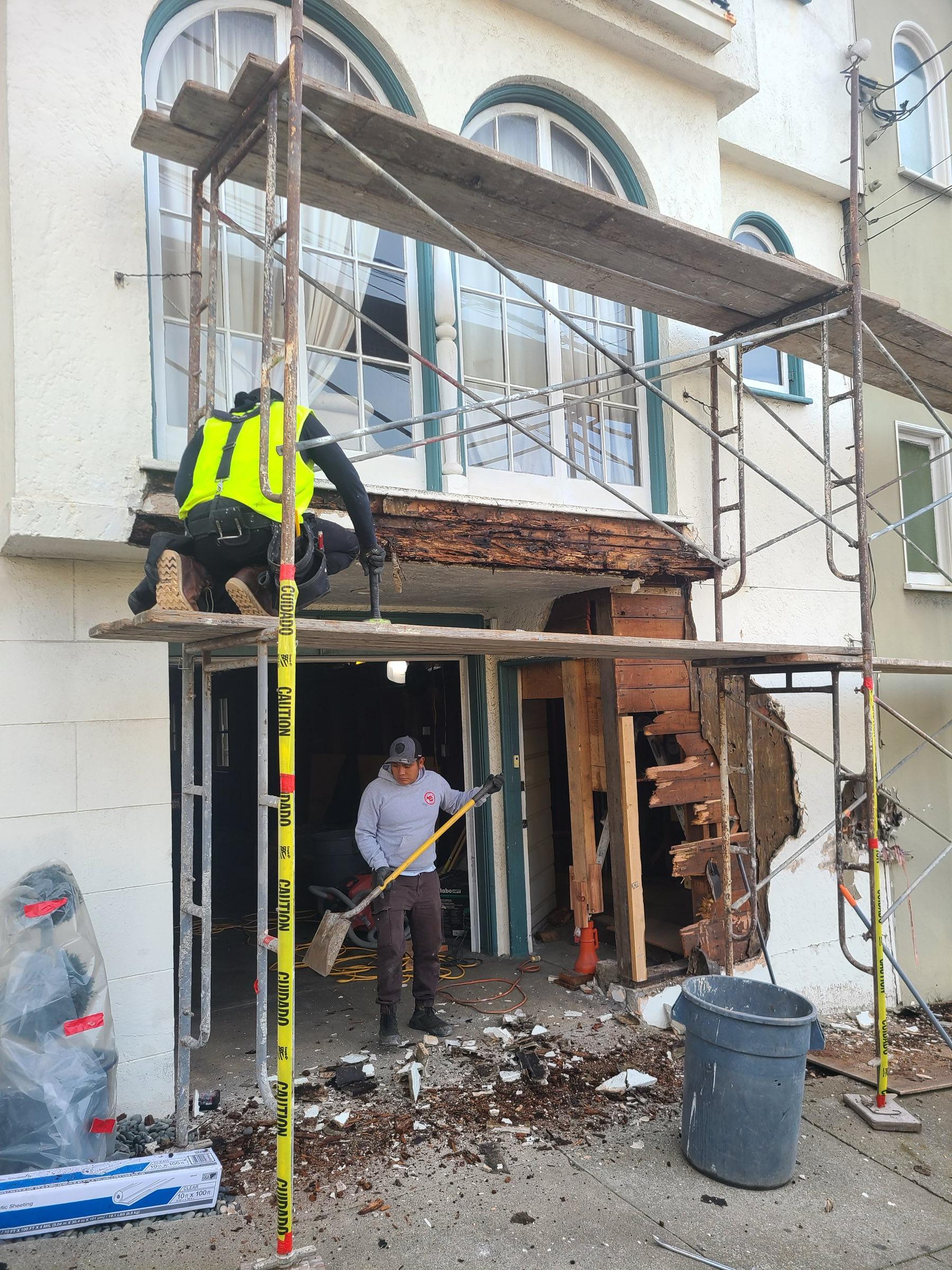 Gadget Construction crew performing dry rot demolition on a San Francisco bay window — exposed rotted framing behind stucco, scaffolding in place, two-man crew with safety equipment
